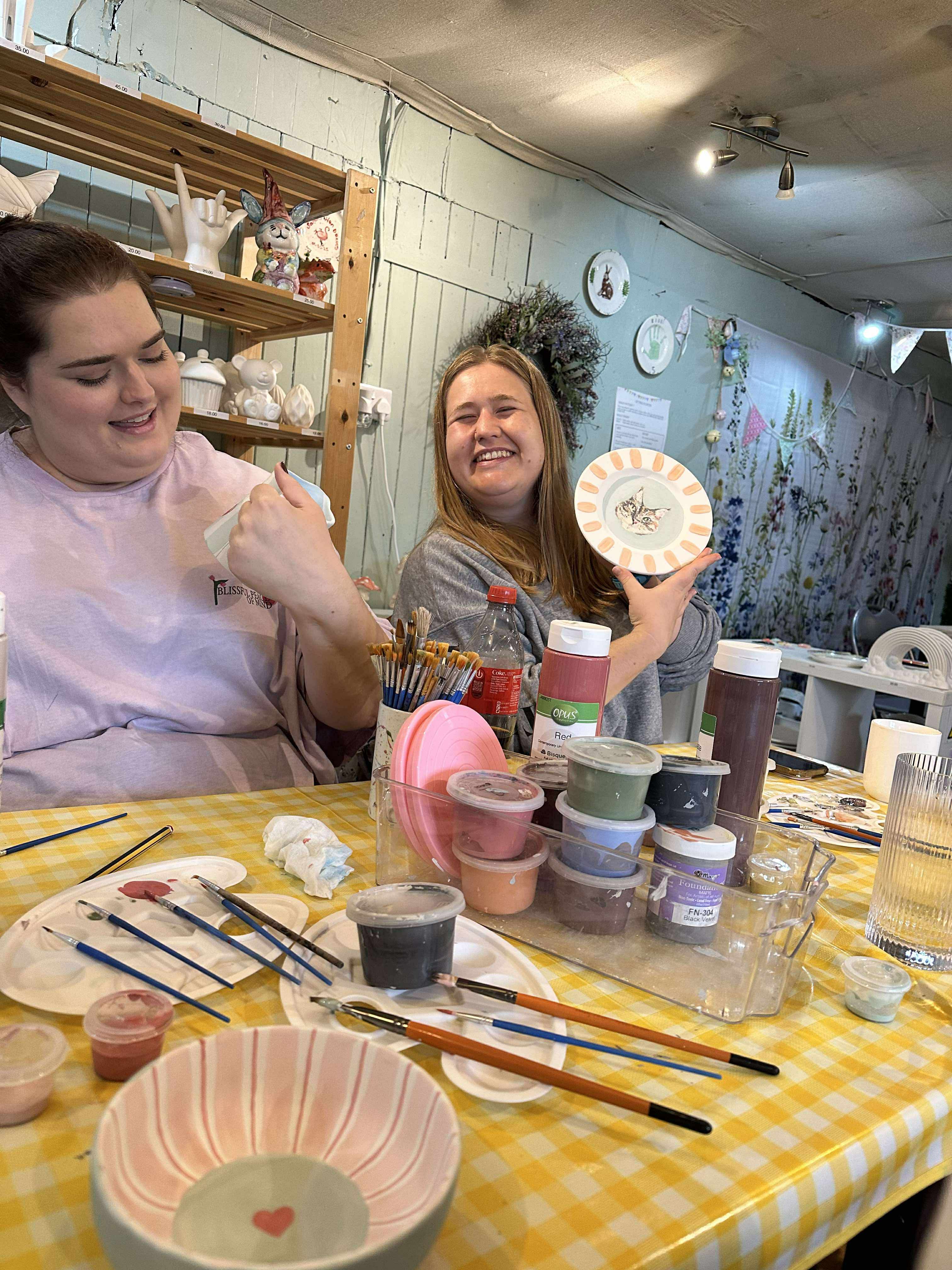 Two friends laughing together at a creative table, one holding a hand-painted plate